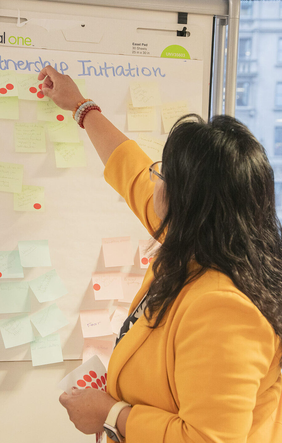 Woman in yellow blazer adds a sticky note to a white board.