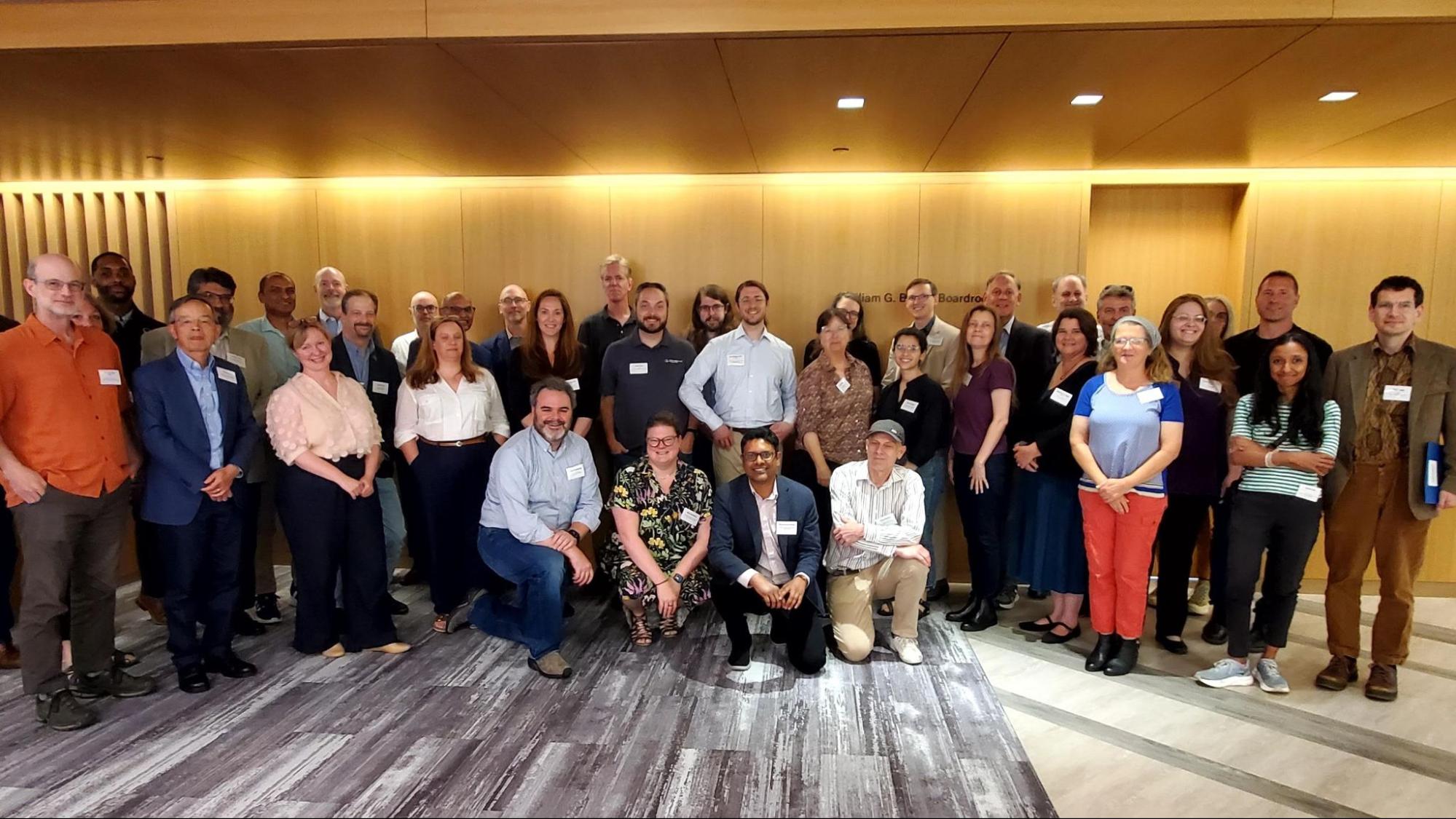 A group of 40 people wearing nametags smiles and poses in Ithaka S+R’s offices in front of a honey wood wall with gray carpet.