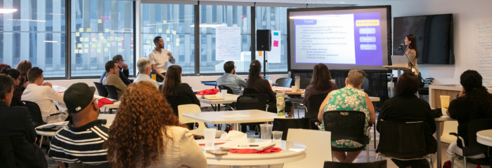A group of people sit around tables in a bright, modern conference room, listening to a presentation. Two presenters stand near a large screen displaying slides, while attendees take notes and engage with the discussion.