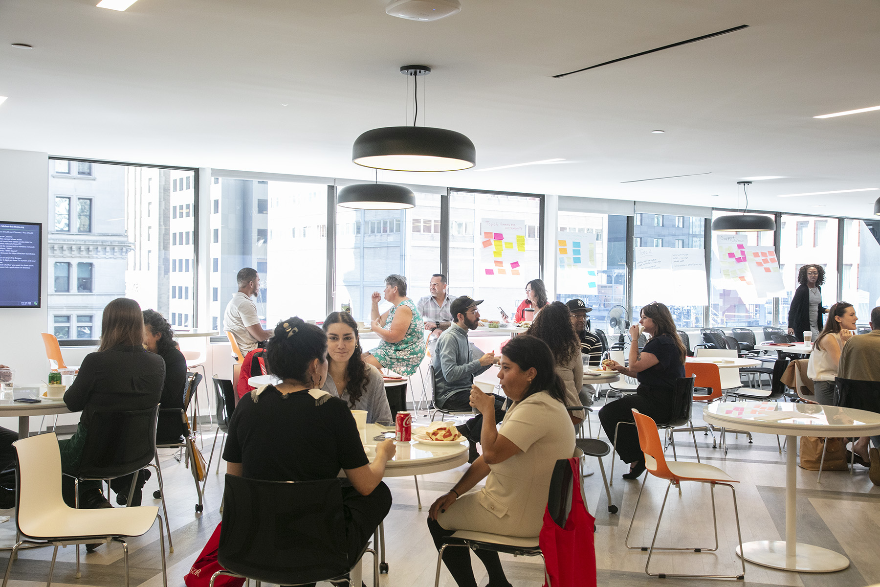 People sit and converse in small groups of two or three at a variety of tables throughout a modern-furnished office. In the background, unreadable sticky notes hang on large sheets of butcher paper, partially obscuring the view of skyscrapers.