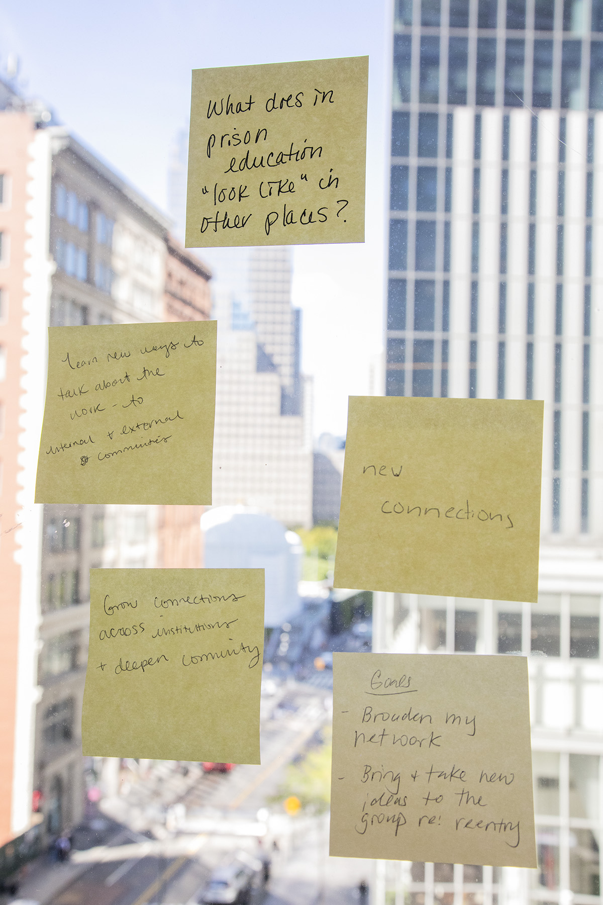 Five sticky notes on a window with New York City out of focus in the background behind them. From top to bottom and left to right, the notes read: “What does in (sic) prison education ‘look like’ in other places?” “Learn new ways to talk about the work – to internal and external communities” “new connections” Grow connections across institutions + deepen community” “Goals - broaden my network -Bring + take new ideas to the group re: reentry”