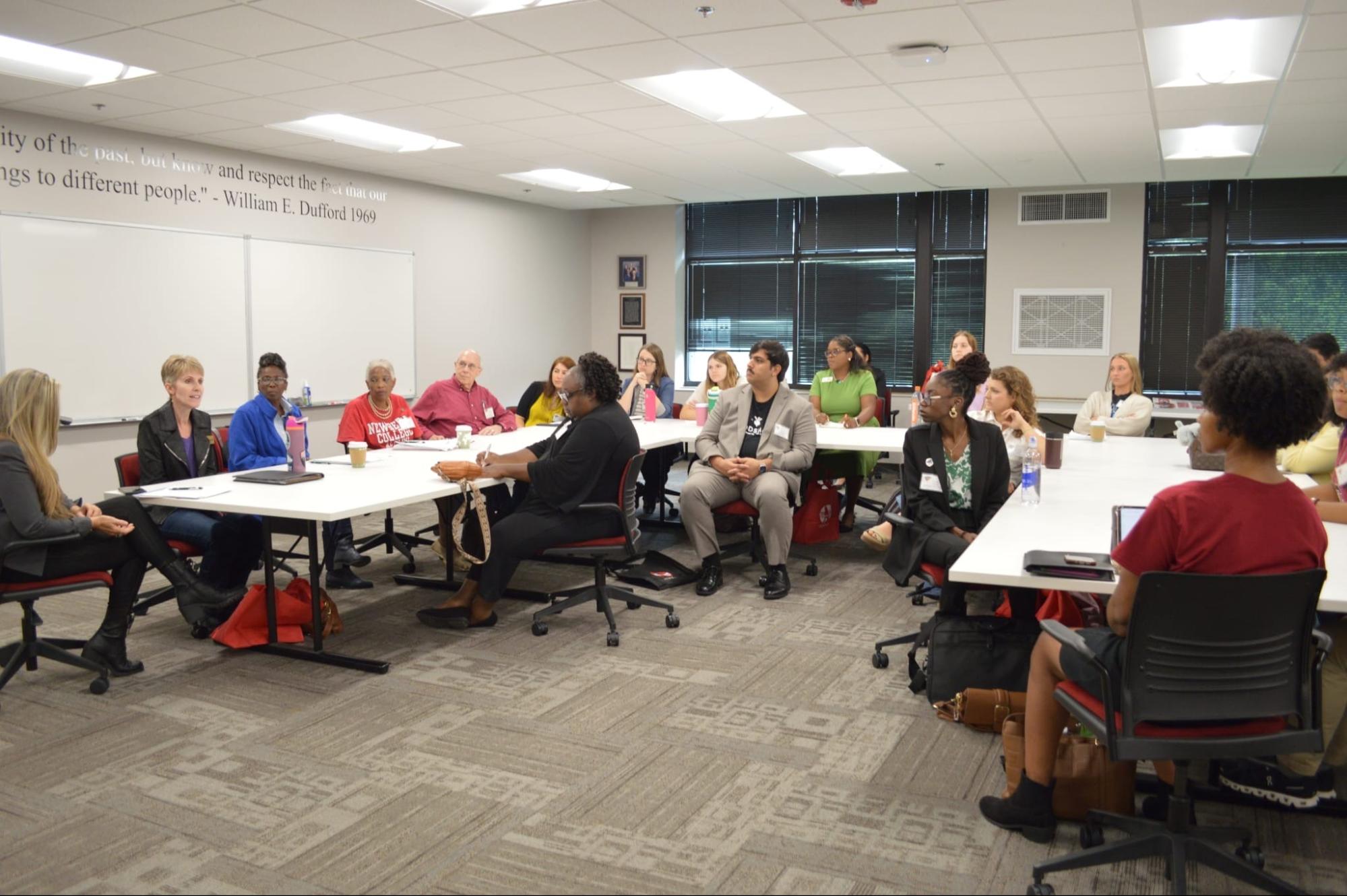 Group of people seated around tables in a meeting room listening to a speaker during a discussion session.