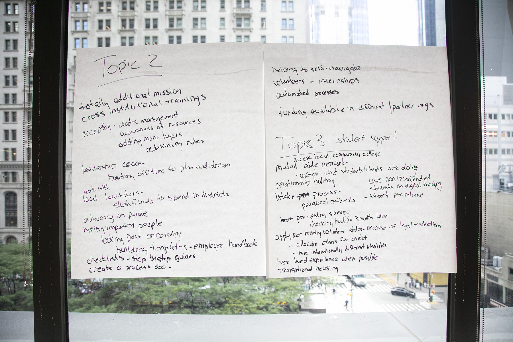 Two large sticky notes made of butcher paper hang from a window side by side. The text on them is difficult to read and appears to be brainstormed lists of solutions to large-scale potential problems that the group identified previously.