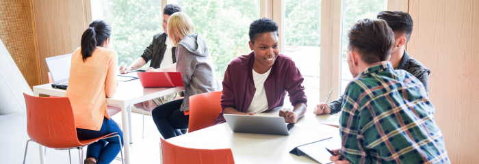 Students in bright college library center collaborate at small tables, with laptops and notebooks.