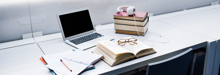 Student's study area in a modern library, including laptop, pile of books, headphones, book and glasses, and notebook and pencil.