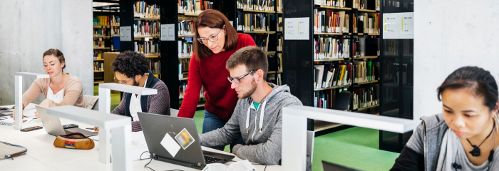 Librarian helps college student sitting with laptop in modern academic library. Behind the student, rows of library shelves with books are visible.