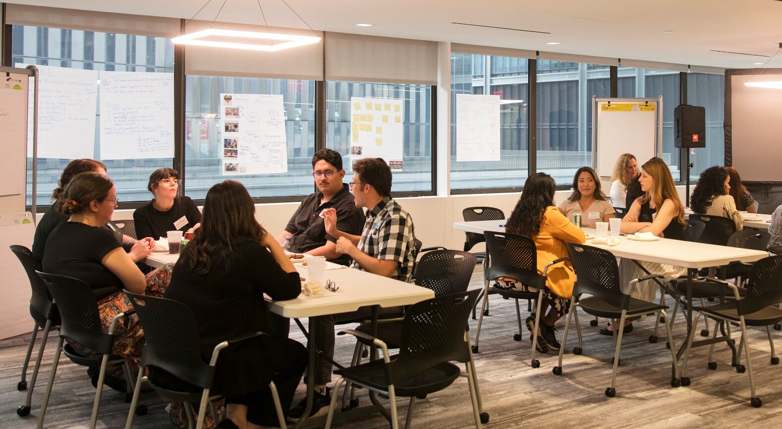 Two small groups of participants sit at tables and brainstorm ideas in a bright office space. On a wall of windows behind them, posters are visible with an array of sticky notes.